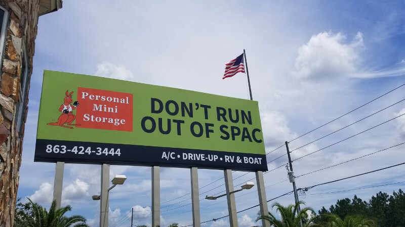 A large billboard with a kangaroo logo reads "DON'T RUN OUT OF SPAC" for Personal Mini Storage. An American flag flutters above, against a partly cloudy sky.
