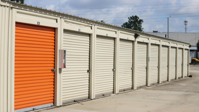 Row of storage units with metal roll-up doors, one painted orange among beige ones. The setting is outdoor with a concrete floor and cloudy sky.
