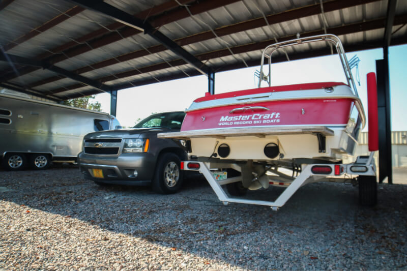 A red and white MasterCraft boat is hitched to a gray SUV under a metal roof. The setting is a gravel-covered storage area, suggesting casual outdoor storage.