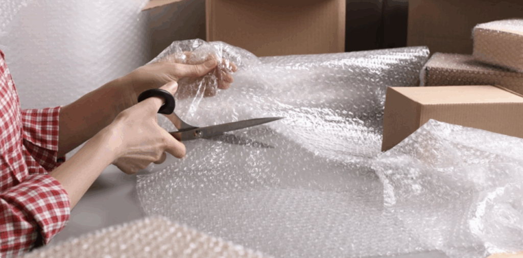 Close-up of a person cutting a sheet of bubble wrap with scissors, surrounded by cardboard boxes, preparing packaging materials for shipping or storage
